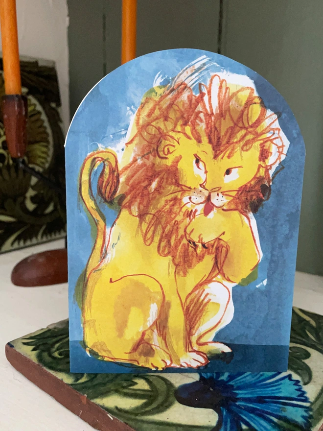 Esther Kent Lion illustrated arch-shaped greeting card, showing yellow and brown lion licking a paw, against a blue background.
