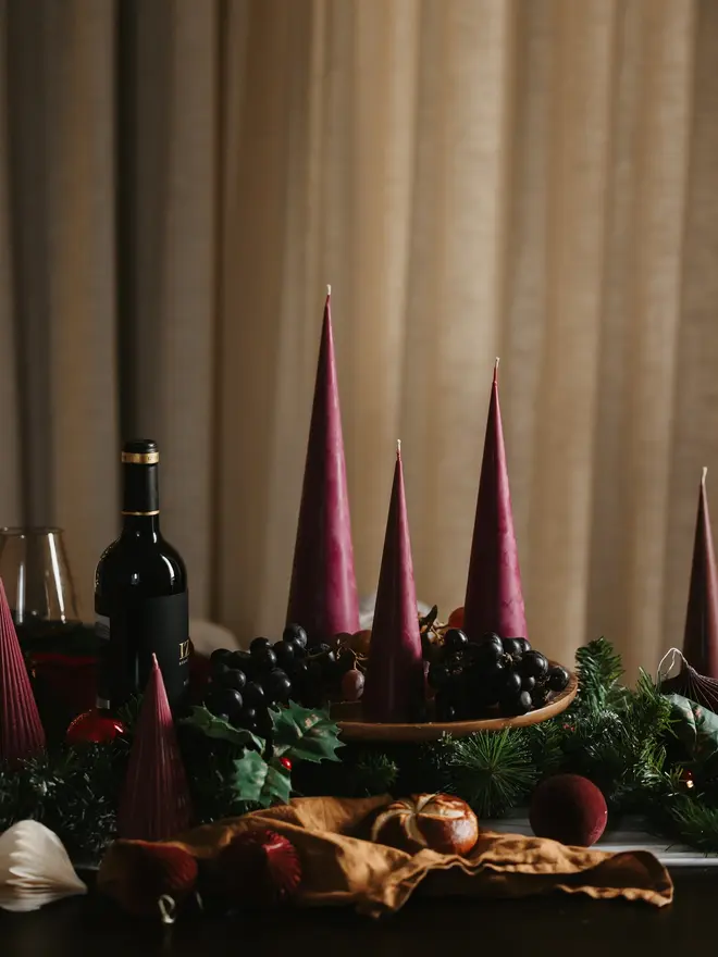 Burgundy Christmas Tree Cone Candles (Set Of 3). A long festive table lined with burgundy cone candles, a bottle of red wine, grapes and pine garland, with fairy lights glowing behind. Tall and short candles together give the tablescape height and drama for holiday entertaining.