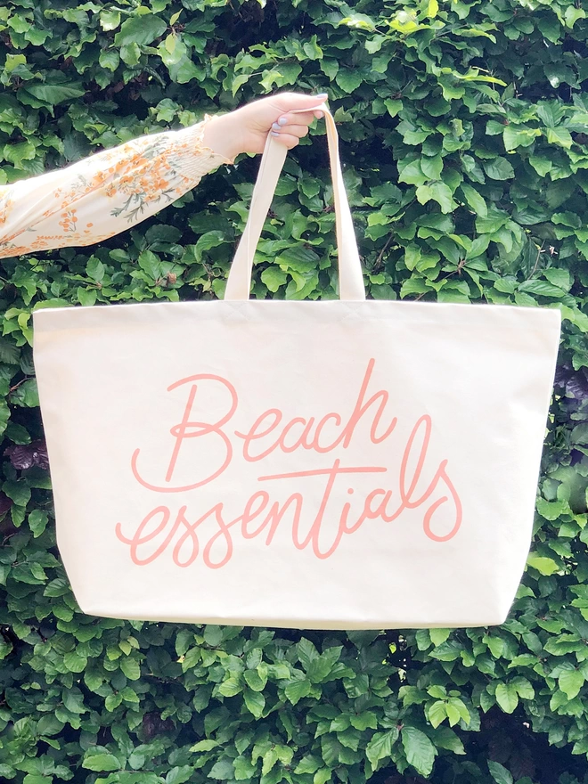 model holding up an oversized natural canvas tote bag with beach essentials slogan