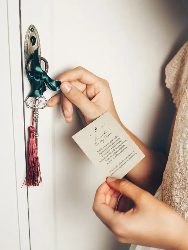 A silver key with a green ribbon and red tassel hangs from an antique door handle. A child holds the "A note for the key keeper" card beside it.