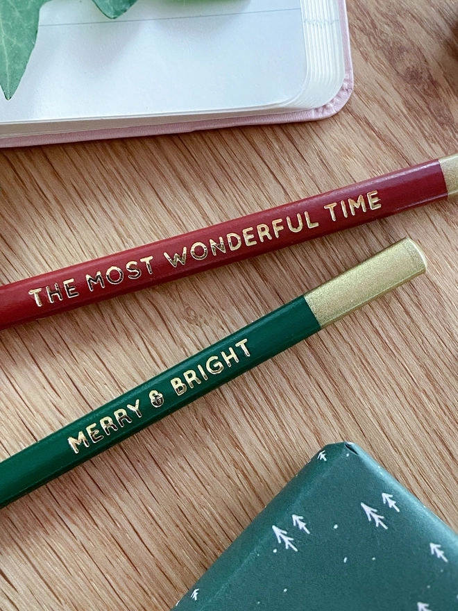 Two pencils, one red and one green, both with gold writing along the sides lay on a wooden desk.