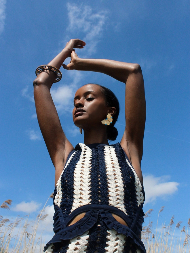 A portrait of a model wearing Nalla Earrings and a Bangle, taken from a lower angle. The model is posing with her arms and hand up in the sky.