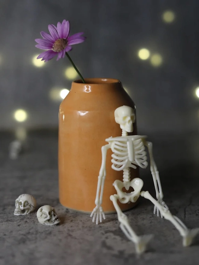 Classic Bud Vase, a colourful ceramic vase sitting on a plain surface against a grey backdrop. It is accompanied by some small skeletons and a flower. 