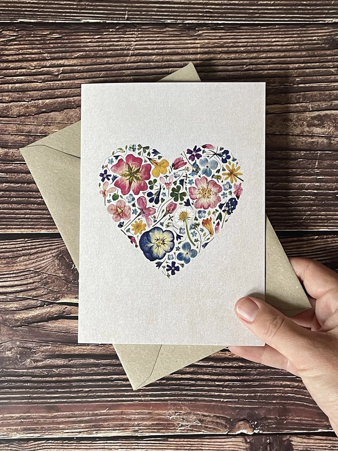Hand Holding Greetings Card with Heart Design Made from Colourful Pressed Flowers - Brown Kraft Envelope - Dark Wooden Background