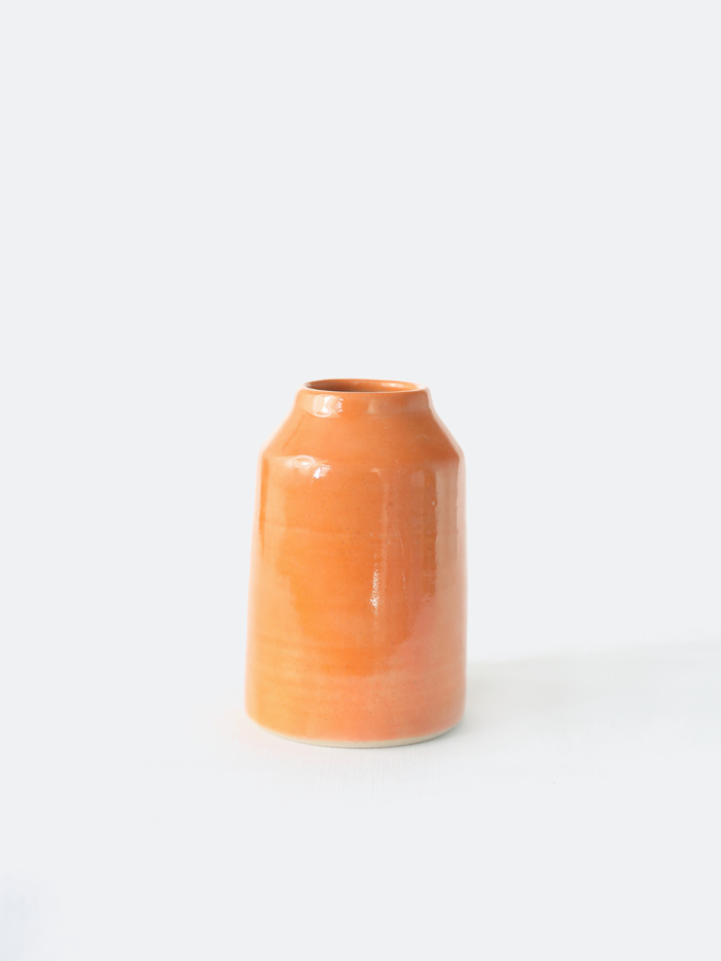 A small handmade ceramic bud vase in glossy mandarin orange glaze, with a rounded bottle shape and narrow neck, photographed against a plain white background