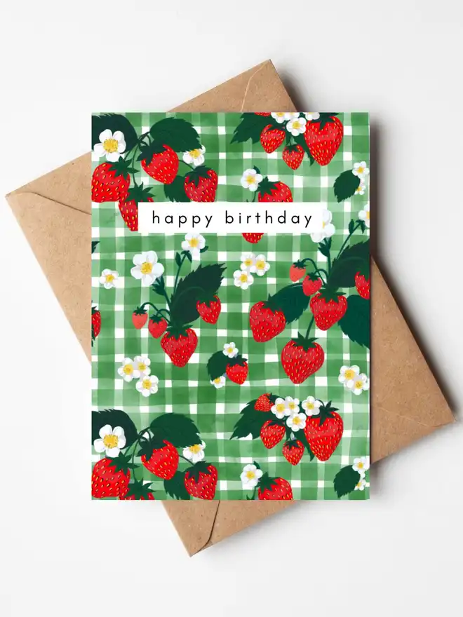 A birthday card featuring hand-illustrated strawberries, strawberry flowers and green leaves on a green gingham background, with the words "happy birthday" across the centre. The card is displayed with a kraft envelope.