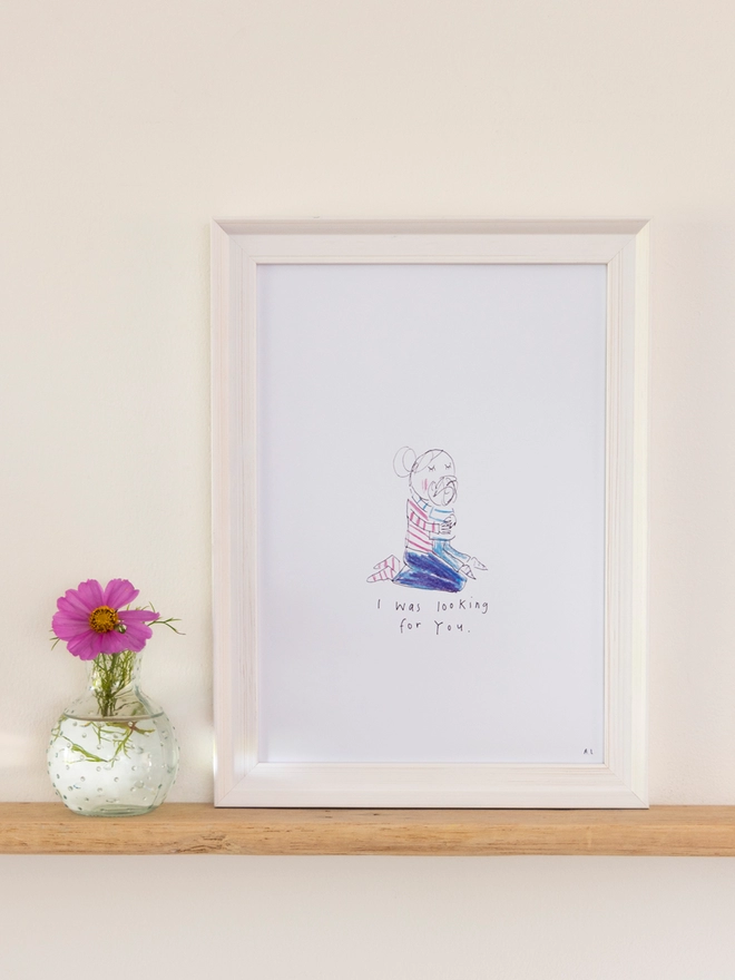 A sketchy muma print in a white frame next to a pink flower in a vase
