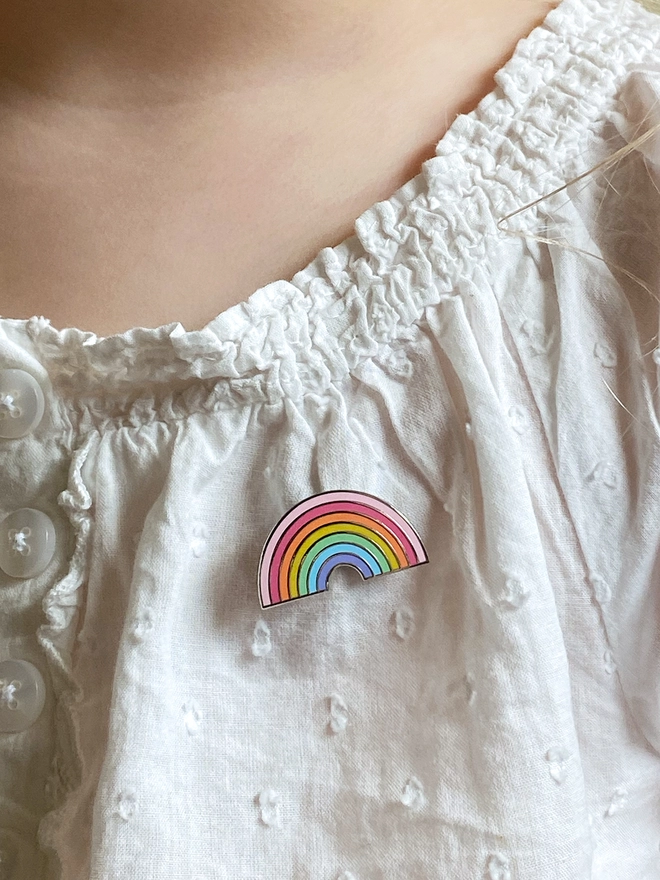 A small pastel rainbow enamel pin badge is pinned on a white top. 