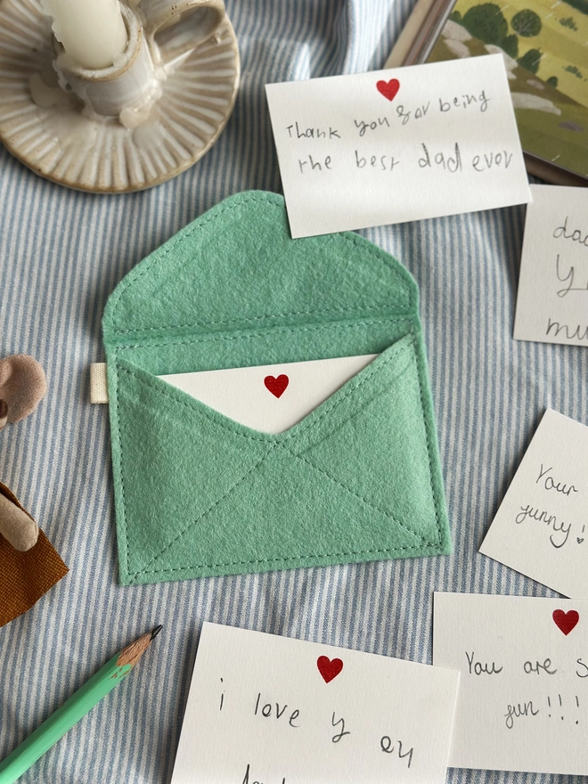 A handmade envelope lays open on light blue striped fabric. Tucked inside are mini heart notecards, with several written cards beside it. Each written card has a note to Daddy written by children.