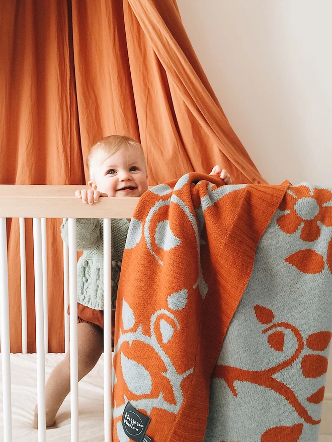 A close up of a smiling baby standing up in a wooden cot. A rust orange canopy creates a curtain in the background, and the rust briar rose junior blanket is draped over the side of the cot in the foreground showing the floral design and reversible colourways. 