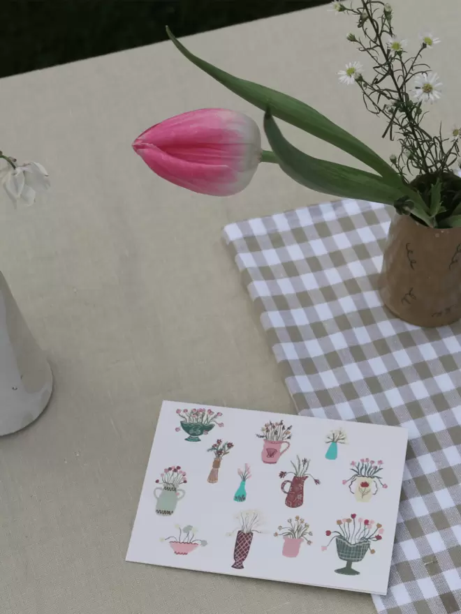 greetings card with floral arrangements on it, with fresh flowers in a vase on a gingham table cloth