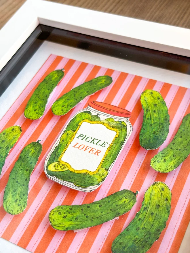 Charming framed collage featuring vibrant illustrated pickles, set against a colorful red and pink striped background, with gherkins and a pickle lover's jar.This vibrant and lovingly crafted mini collage is super cute and will delight pickle lovers and gherkin fans alike!