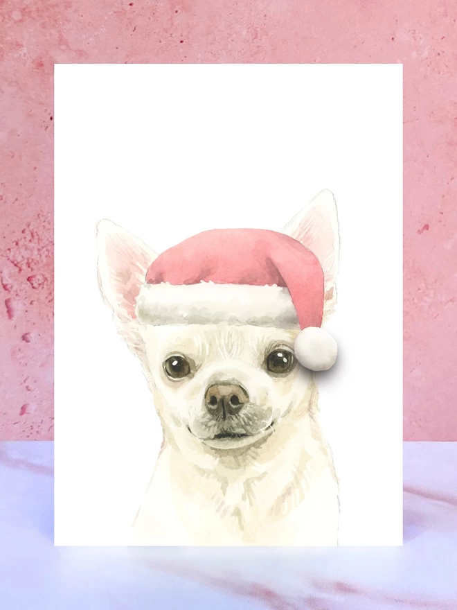 A Christmas card featuring a hand painted design of a chihuahua, stood upright on a marble surface surrounded by pompoms. 