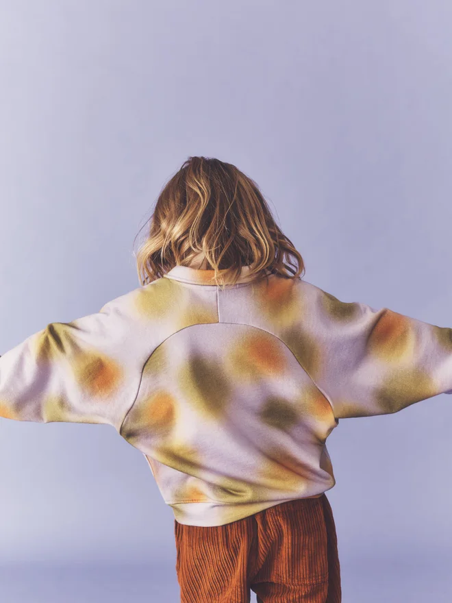 Back view of a child with arms outstretched, showing the curved back yoke of the Studio Koter Luka jersey top in Hazy Lilac, worn with Ever trousers in umber cord.