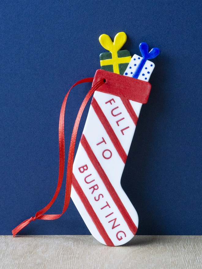Ceramic porcelain Christmas decoration with Full to Bursting written on the side. A white stocking-shaped Christmas decoration with red lettering, decorated with red stripes and with a red satin ribbon.