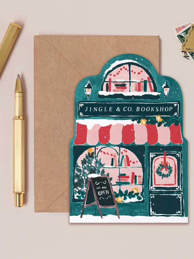 Teal Bookstore Christmas card shaped like the Jingle & Co. Bookshop front with a striped canopy and snowy roof, on a kraft envelope beside a gold pen