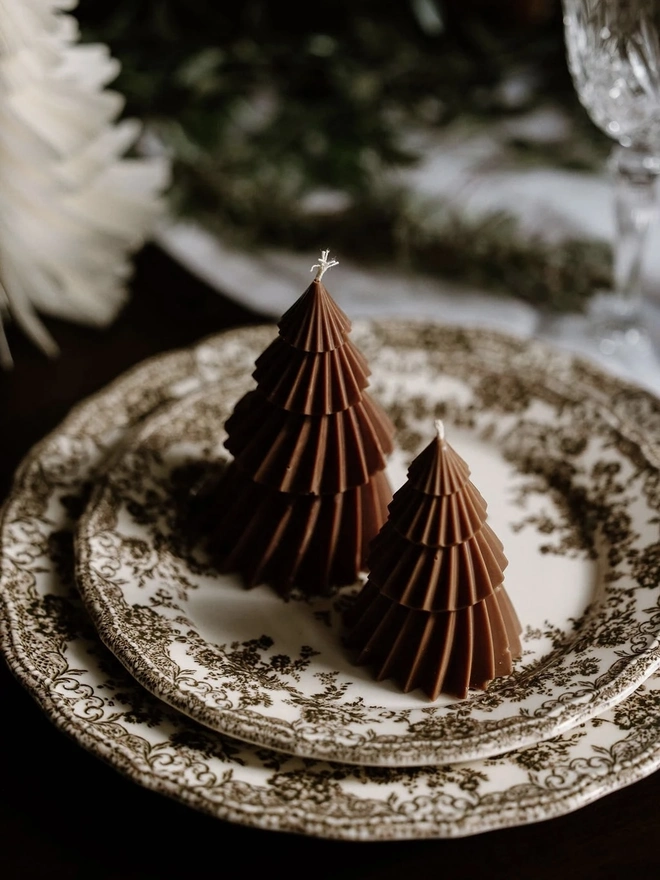 Brown Christmas Tree Warm Candles (Set Of 2). Two chocolate brown Christmas tree candles with fluted, layered tiers sitting on a vintage brown floral transfer-print plate. The rich earthy tone makes a stylish place setting decoration for Christmas dinner.