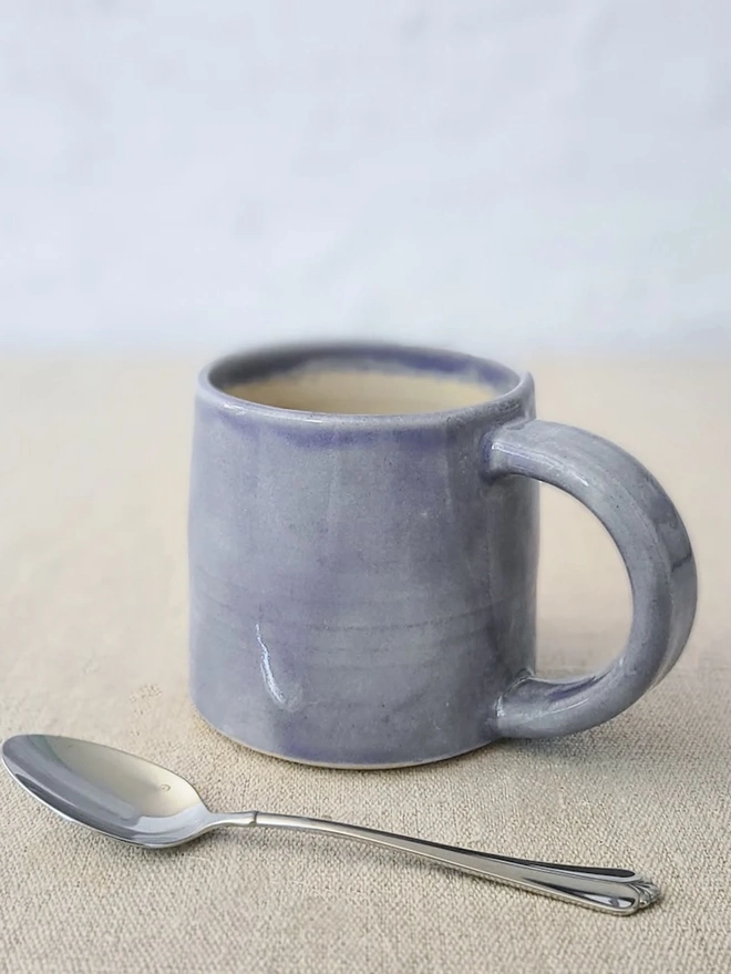 Ceramic Standard Mug, a colourful ceramic mug sitting on a plain surface against a grey backdrop. It is accompanied by a spoon. 