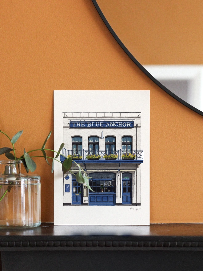 An architectural illustration of The Blue Anchor pub on a print, displayed on a dark mantle next to a glass vase of eucalyptus against a mustard wall.