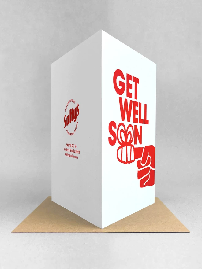 Rear view of the get well soon card, design shows a finger wrapped in a bandage form the two o’s of the word SOON. Printed in red ink on a white portrait format card. Stood on a kraft brown envelope, in a light grey studio set.