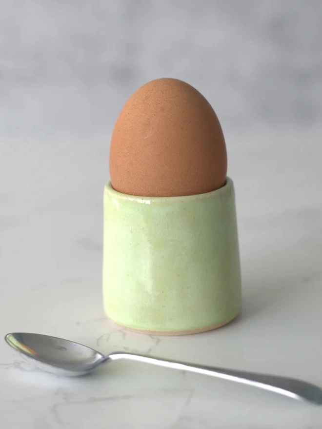 Classic Handmade Egg Cup, a ceramic egg cup sitting on a plain surface against a grey backdrop. It is accompanied by a spoon. 