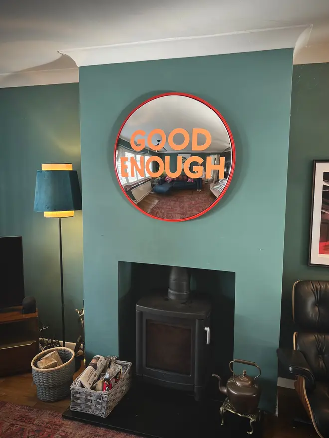 Round convex mirror with 'Good Enough' hand painted in orange in a red round frame, on a teal wall and log burner, with a lamp, logs and white ceiling