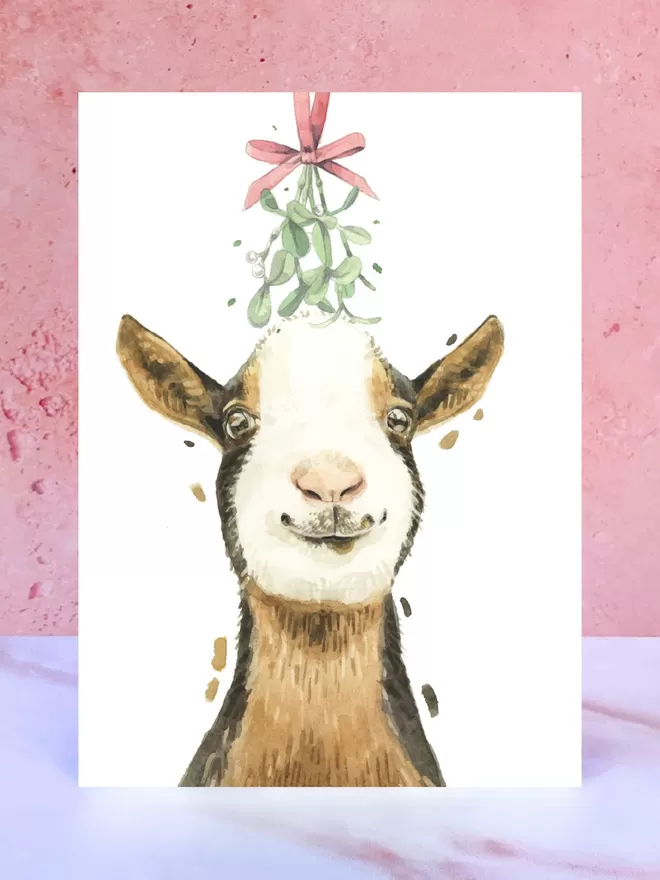 A Christmas card featuring a hand painted design of a Goat, stood upright on a marble surface.