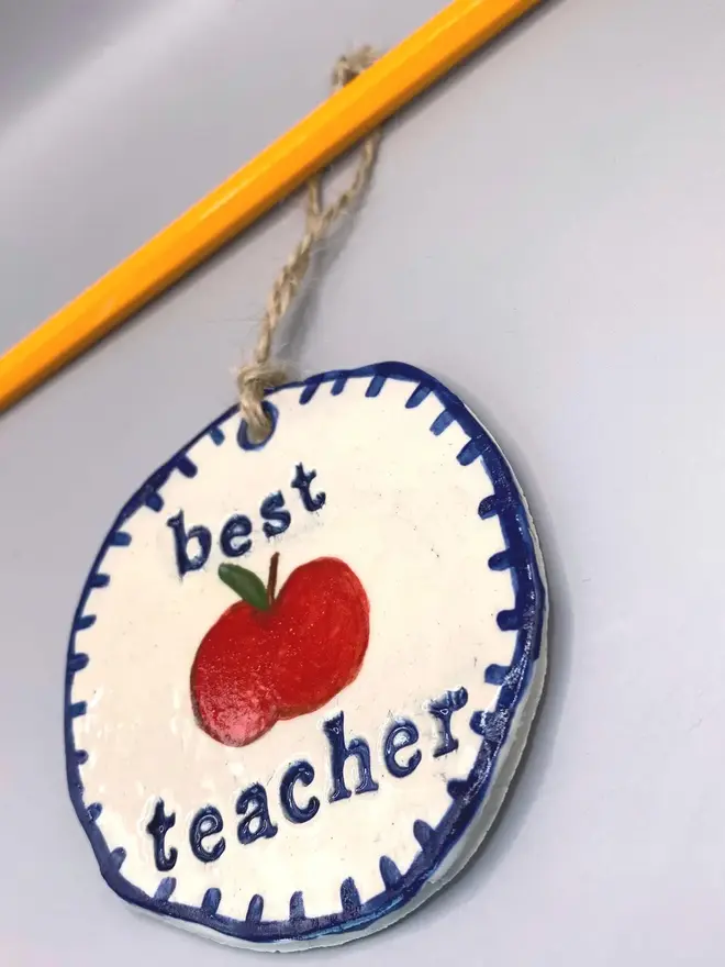 'Best Teacher' Handcrafted Ceramic Ornament, a ceramic apple ornament sitting on a plain white surface hanging from a pencil. 