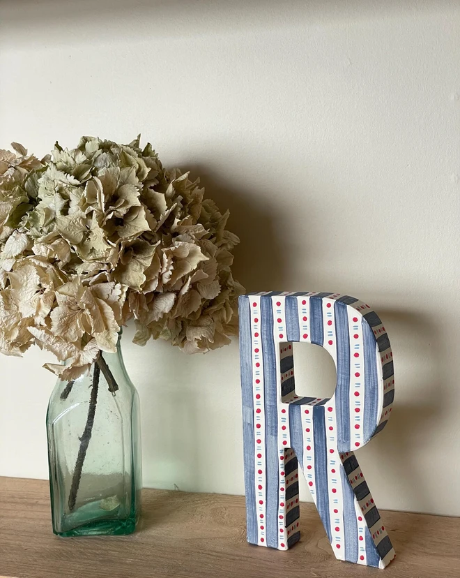A hand-painted decorative initial letter  with stripes on a wooden table against a light wall. 