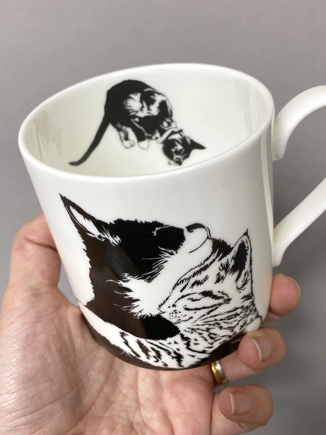 Cat mug, showing two cuddling cats on the front, angled so you can see a black cat leaping down inside.