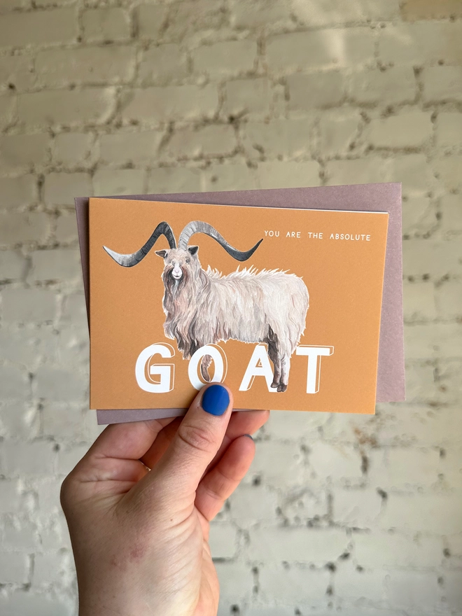 an orangey yellow greetings card featuring an illustration of a bilberry goat with the text “you are the absolute GOAT” with a muted purple envelope