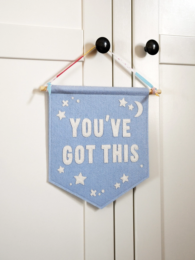 A blue felt wall hanging, with the words You’ve Got This and white felt stars, hangs on a white wardrobe from its ribbon hanger.
