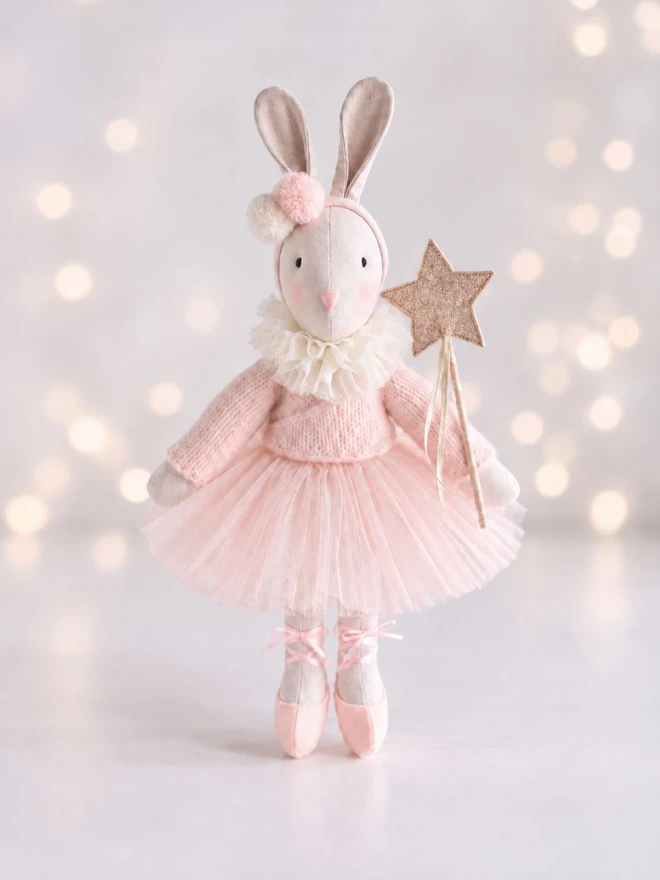 Verity ballerina bunny standing with a gold star wand against a soft pale background with glowing bokeh lights.