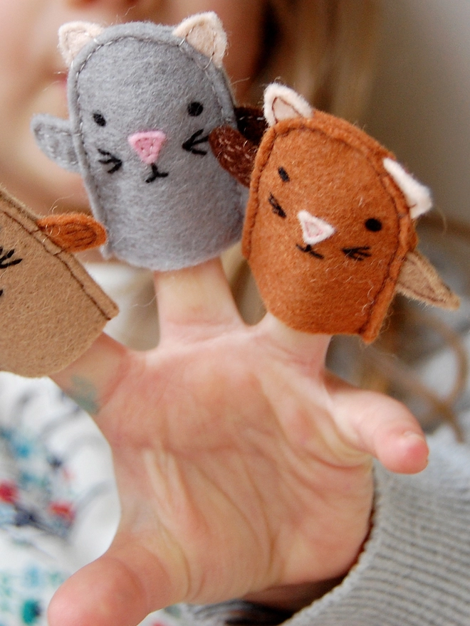 A young child holds three homemade felt kitten finger puppets in her hands.