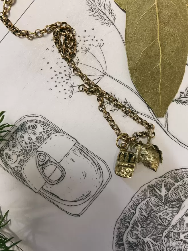 Image of hand carved gold toned brass charms hanging from chain. One charm is a tin of sardines and the other a small beetroot. Laid on illustrated calendar at background showing sardines, fennel and cabbage etchings. 