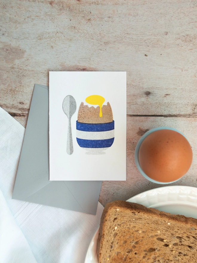 A cute little greeting card with an illustrated boiled egg in a blue and white striped egg cup rests on the breakfast table with a boiled egg and hot toast nearby.