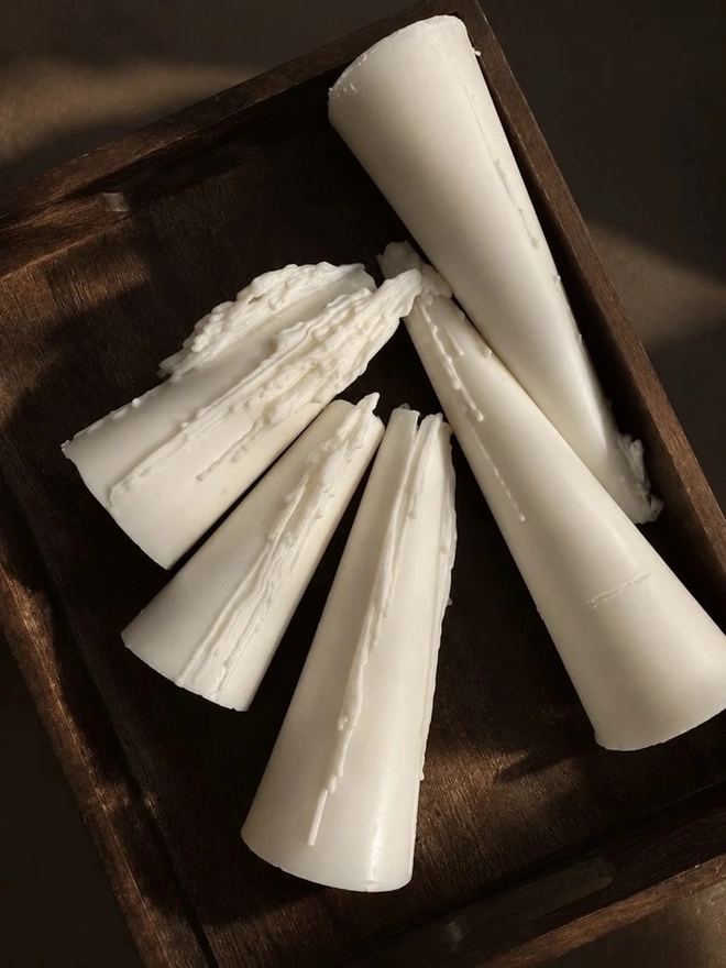 White Christmas Tree Cone Candles (Set Of 3). A top-down view of cream cone candles with sculpted wax drips, laid in a dark wooden tray in soft sunlight. The textured drip detail gives each handmade candle its own unique character.