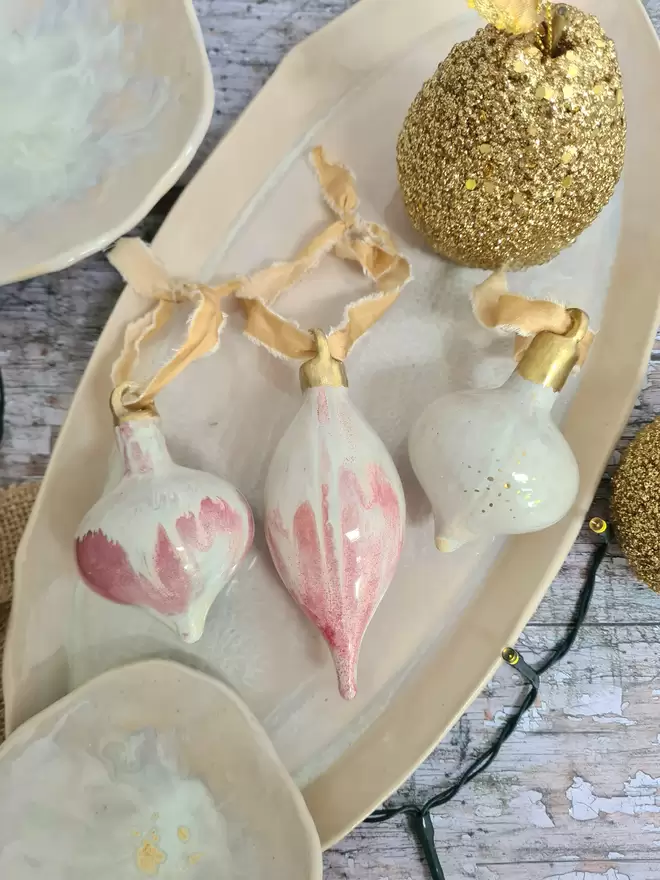 3 Ceramic christmas bauble decorations in a pearlescent glaze with white, cream and pink and gold, with silk ribbon and gold detailing. Photographed on a pearlescent serving dish with gold pears and christmas lights