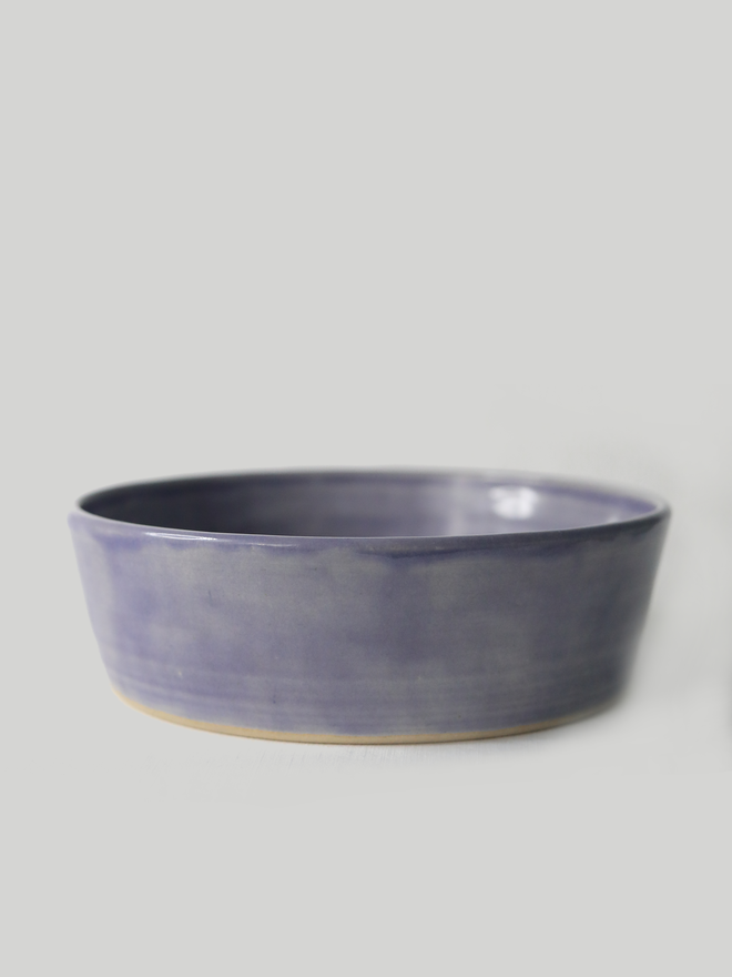 A single wide, shallow glazed ceramic pasta bowl in amethyst purple, photographed from a slight angle against a plain light grey background, showing the unglazed natural clay base.