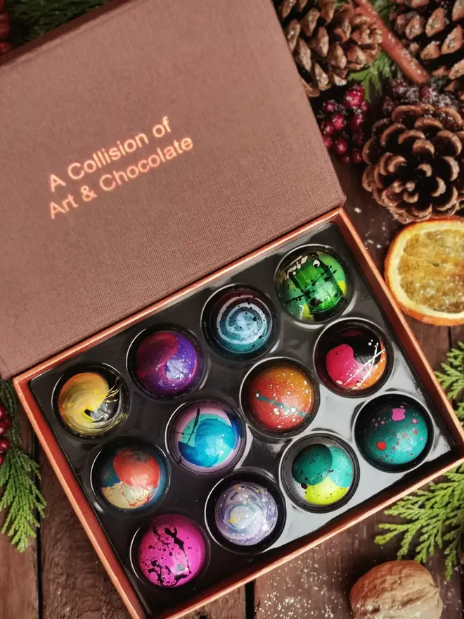 box of 12 beautifully decorated, colourful chocolates, on a background with pine cones and oranges