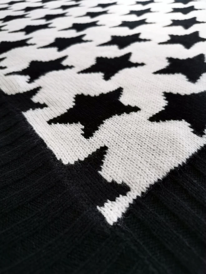 Close up of the corner of a knitted blanket showing the detail of the ribbed trim, the knit stitches and black and white star pattern.