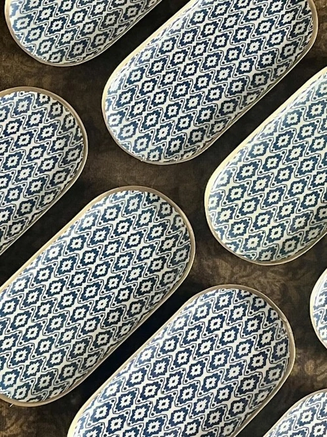 Perfect Enamel Oval Tray. A collection of navy and cream ikat-patterned oval trays with gold rims laid out on a dark textured surface. The repeating geometric design shows off a coordinated set of decorative trays for the home.