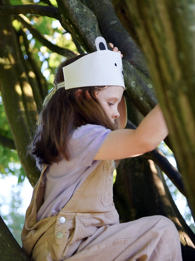 A young girl wearing a dress-up polar bear fabric crown and beige dungarees is climbing a tree.