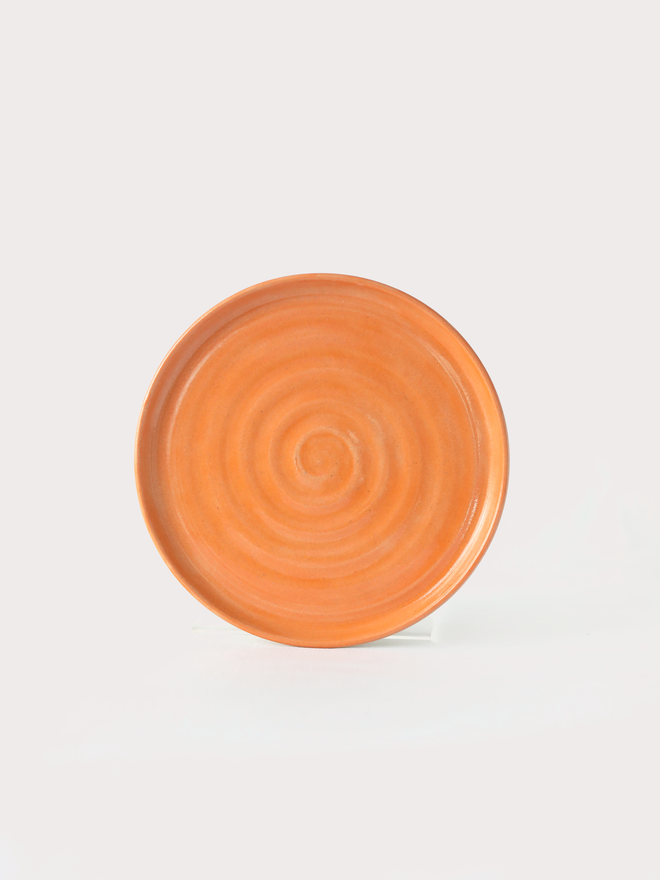 A handmade ceramic cake plate in mandarin orange glaze, photographed from the front against a plain light grey background, showing a natural concentric swirl texture and a shallow raised rim