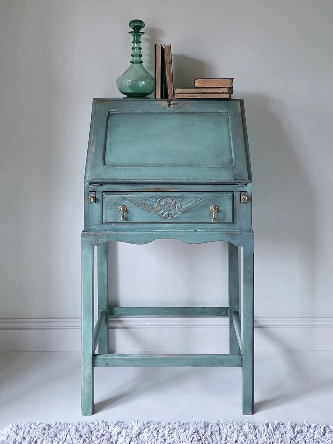 Vintage 1930s Oak bureau with a single drawer and carved floral detail on the front. It has original brass drop handles staged with vintage Green decanter and antique books rested on top.