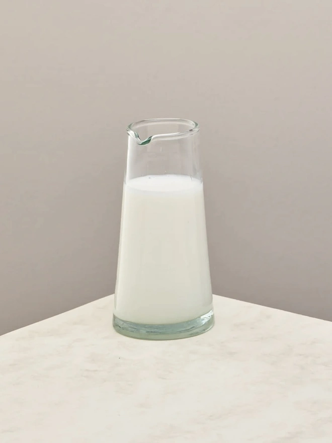 Tall Jug, a tall glass jug sitting on a plain counter top against a white backdrop. It is filled by milk.