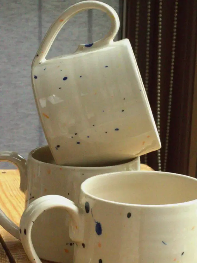 Two large creamy white terrazzo cups with black, blue, and yellow splatters and glossy glaze stacked on each other on a wooden surface.