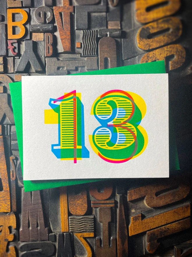 Colourful handmade 13th birthday letterpress card paired with a contrasting green GF Smith Colorplan envelope. The bold number 13 is printed in overlapping cyan, magenta and yellow inks on luxury Pristine White card and photographed against vintage wooden letterpress type.
