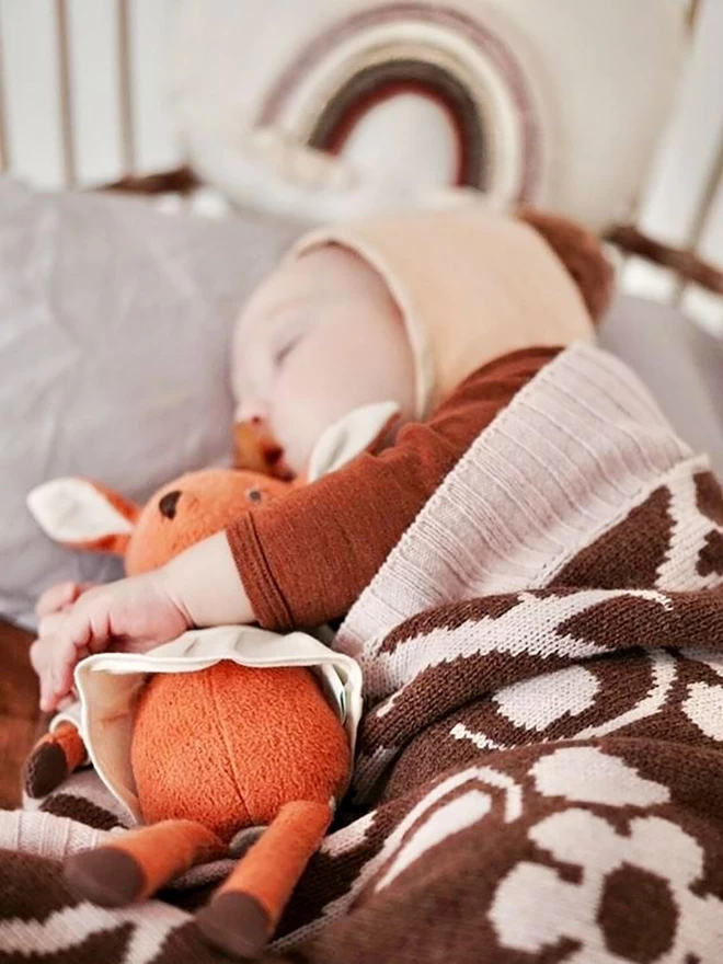 A baby lies asleep in its cothugging a brown fawn doll. The baby is cuddled up in the brown knitted blanket with pink flower design. A matching coloured rainbow hangs in the background.