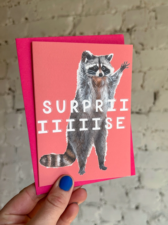 a bright coral pink greetings card featuring an illustration of a raccoon with its hands in the air with the text “surpriiiiise” with a neon pink envelope